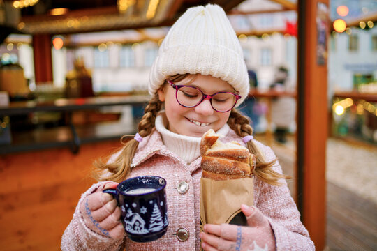 Little cute school girl eating sweet bread called Baumstriezel and drink hot chocolate or punch on German Christmas market. Happy child on traditional family market in Germany,