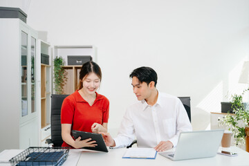 Two Asian coworkers collaborating on business planning, discussing documents and using digital devices in modern office