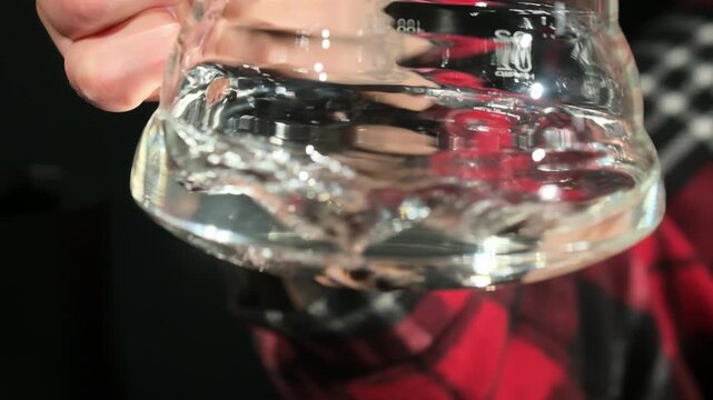 Hand tilts glass measuring cup causing liquid movement. Person shakes transparent jar with volume markings creating sloshing motion slow motion