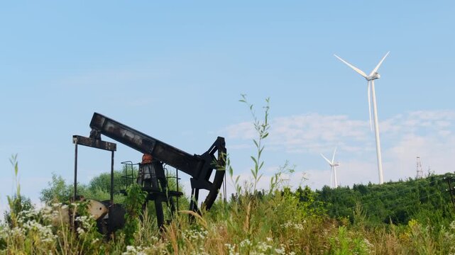Oil pumpjack dips rising beside tall wind turbine on ridge. Steel beam swings with rod pumping slowly while grassy field stretches under blue sky