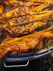 A close-up vertical shot of glazed, whole grilled chickens laid flat on a metal grate over hot charcoal, with thick bamboo sticks holding them in place for cooking.