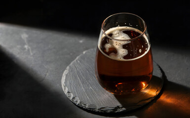 Moody amber beer glass with foam on slate coaster, dark background with dramatic light and shadow, rustic pub drink, refreshing cold alcoholic beverage, elegant still life photography, closeup liquid.