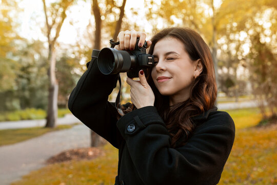 Young woman reviewing photos on her digital camera while standing in a sunny park. Warm autumn light, creative outdoor photography moment, lifestyle scene.
