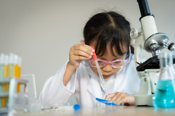 A young girl is wearing a lab coat and glasses while conducting an experiment. She is holding a red...