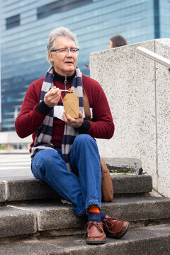 Senior man sitting on steps in urban plaza eating from takeout container with chopsticks