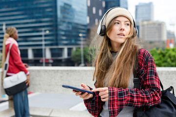 Diverse women standing on city terrace railing holding phone and wearing headphones, copy space