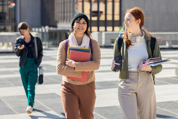 Female student friends walking across campus plaza carrying notebooks and checking phones