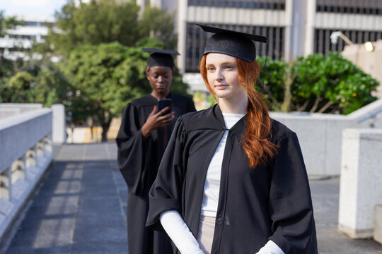 Diverse female graduates wearing gowns and mortarboards, gazing and scrolling smartphone on terrace