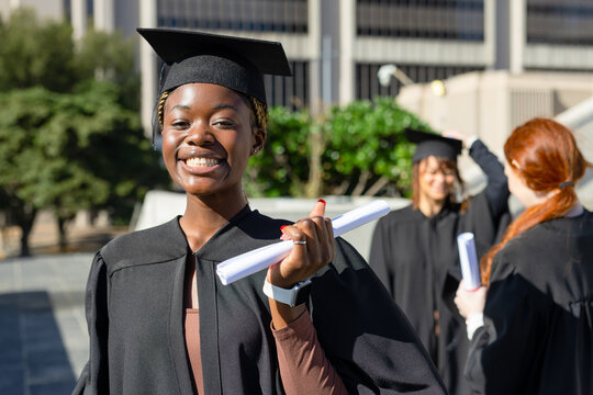Diverse classmates standing on plaza wearing black gowns and mortarboards holding rolled diplomas