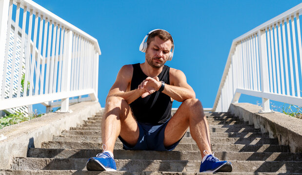 Male runner checking fitness tracker while resting on urban steps after workout.