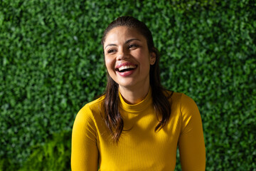 Mid adult woman wearing mustard-yellow top standing against green hedge wall and smiling outdoors