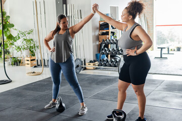 Diverse female workout partners high-fiving on rubber mats near kettlebells and battle ropes