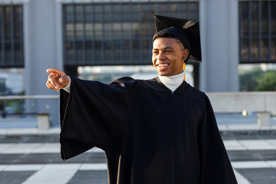African American man wearing graduation gown and mortarboard cap pointing left on rooftop plaza