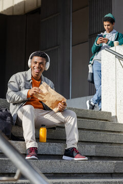 African American classmates sitting campus steps eating from paper bag and scrolling smartphone
