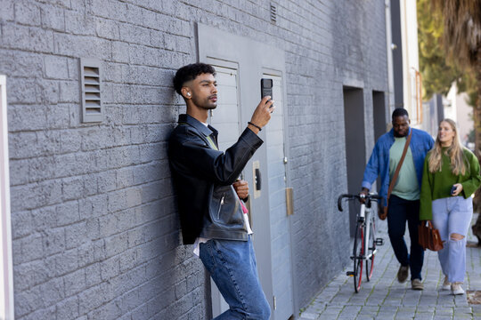Man using smartphone, using earbuds, leaning by utility door, couple pushing bicycle with brown bag