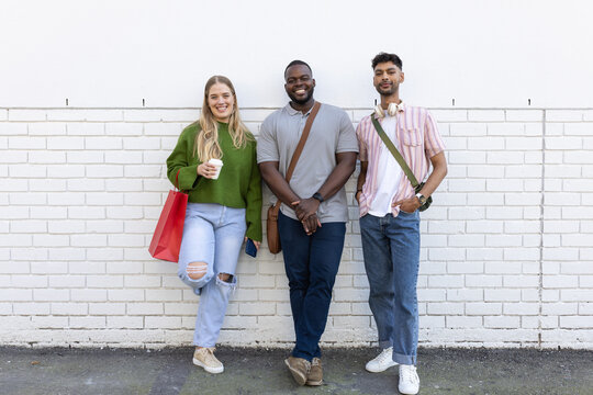 Diverse friends leaning on wall holding coffee cup, red bag, smartphone, messenger bag, headphones - Powered by Adobe
