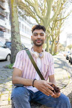 Indian male sitting on ledge on tree-lined sidewalk eating muffin with headphones and crossbody bag