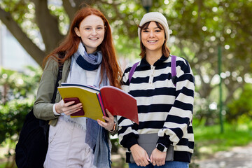 Diverse female students studying on campus walkway holding folder and tablet, wearing backpacks