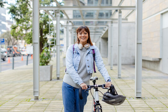 Woman smiling holding bicycle wearing headphones and backpack under metal pergola on walkway - Powered by Adobe