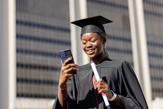 African American woman in gown and cap holding diploma, scrolling smartphone on campus, copy space