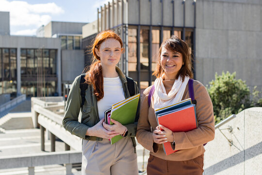 Diverse female friends standing together holding notebooks folders and backpacks on campus walkway