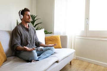 Young man meditating on the couch listening to guided meditation using wireless headphones. Copy space
