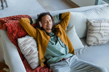 Mid adult man reclining on light gray sofa in living room, wearing mustard jacket, pink headphones