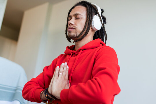 African American man praying with eyes closed in living room wearing white headphones red hoodie
