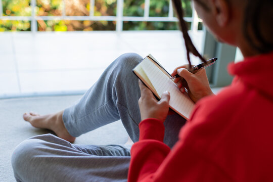 Person writing in notebook with black pen by glass window, wearing red sweatshirt and grey sweats