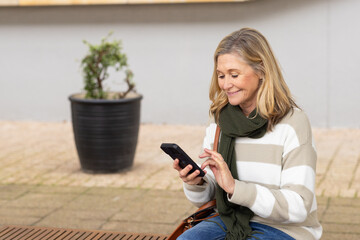 Senior woman tapping smartphone while holding leather handbag on wood bench in plaza, copy space