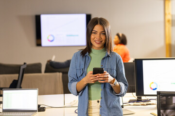 Diverse female coworkers smiling and holding smartphone at open-plan office with chart display