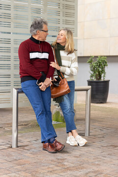 Senior couple leaning on steel rail in urban plaza, checking smartphone while holding handbag