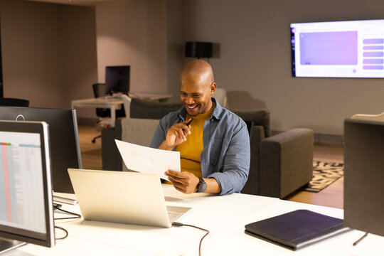 African American man reviewing document at office desk while viewing spreadsheet on laptop monitor