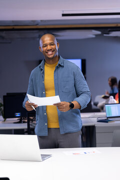 African American man standing at desk holding document in open-plan office with laptop and monitors