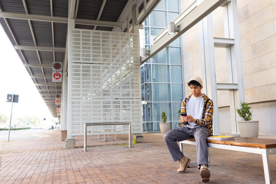 Asian man sitting on wooden bench at transit stop using smartphone with backpack, copy space