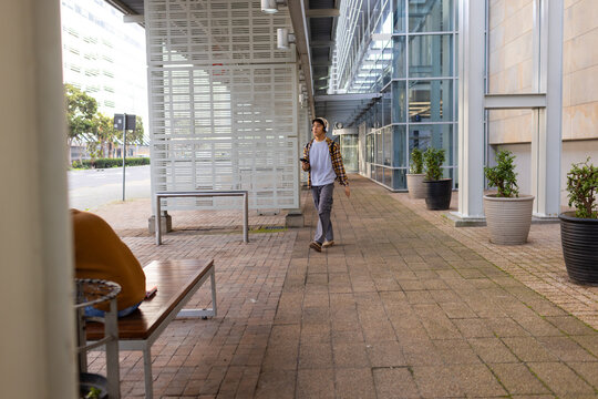 Asian man walking along brick walkway wearing plaid shirt and headphones holding smartphone