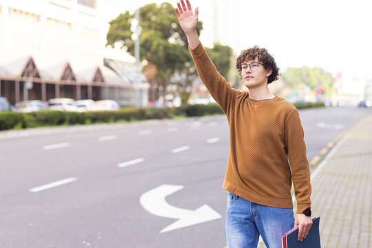 Male student raising right hand at city street while holding blue binder on sidewalk, copy space