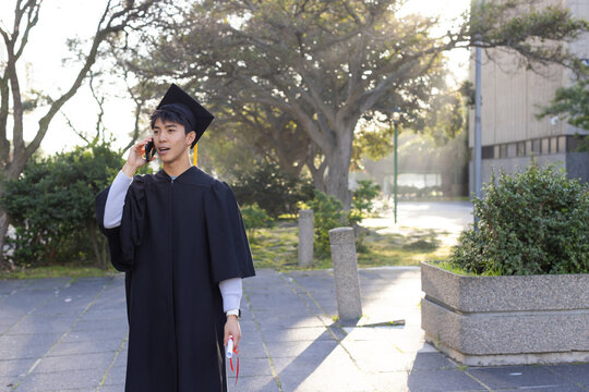 Asian man graduate in gown and cap talking on smartphone holding diploma scroll on campus path