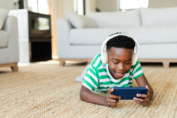 African American boy lying on beige rug at home holding smartphone, wearing white headphones