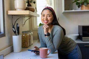 Asian woman smiling and leaning on marble countertop holding smartphone in kitchen with pink mug