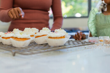 Metal rack holding six vanilla cupcakes on kitchen counter near window, hand adding pastel pearls