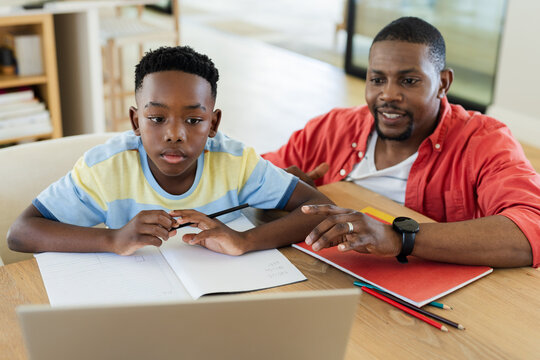African American father and school-age son studying at wooden table in study with laptop notebook