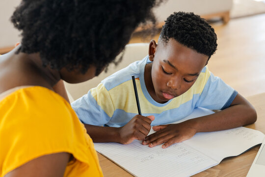 African American mother guiding son writing in notebook at wooden table with laptop at home