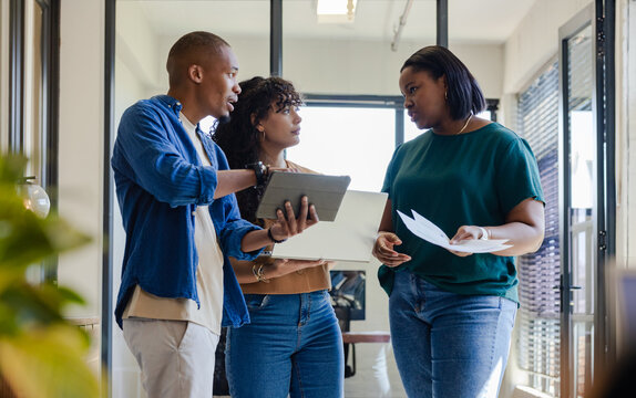 Diverse coworkers reviewing documents with tablet, laptop in office corridor with glass partitions