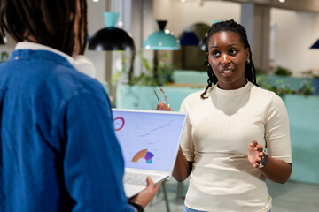 African American coworkers discussing laptop charts while woman holding eyeglasses in open office