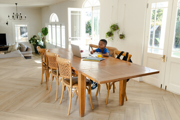 African American boy sitting at wooden dining table working on laptop and notebooks, copy space