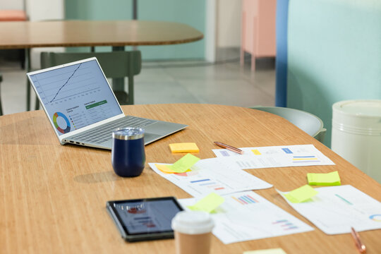 Wooden oval table is holding silver laptop, printed charts, sticky notes, cups in office