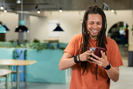 African American man in orange tee smiling, checking red phone and holding metal tumbler in cafe
