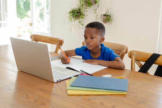 African American boy writing notebook at dining table by window using laptop orange pencil eraser
