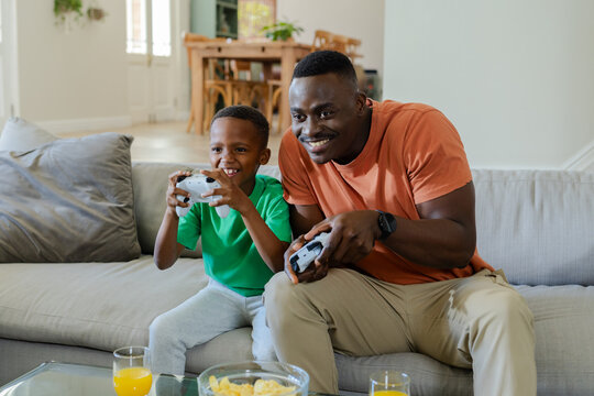 Mid adult father and child son playing controllers on light couch near glass coffee table